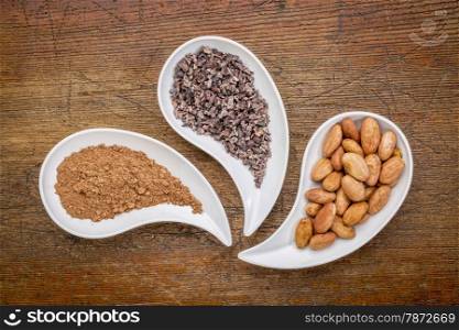 cacao beans, nibs and powder - top view of teardrop shaped bowls against rustic wood