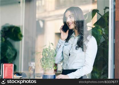 Cafe city lifestyle. Young woman sitting indoor in trendy urban cafe talking with her mobile phone. Cool young modern caucasian female model in her 20s