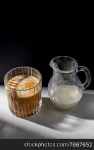 caffeine, objects and drinks concept - glass of coffee and jug with milk or cream on table. coffee in glass and jug of milk or cream on table