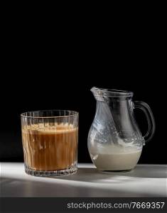caffeine, objects and drinks concept - glass of coffee and jug with milk or cream on table. coffee in glass and jug of milk or cream on table