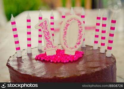 Cake decoration in the figure of the number 10, made of sugar dough and candy in white and pink.The decoration placed on brown chocolate cake.There are also 10 candles on the cake.