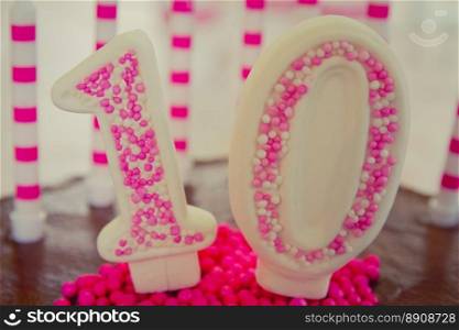 Cake decoration in the figure of the number 10, made of sugar dough and candy in white and pink.The decoration placed on brown chocolate cake.There are also 10 candles on the cake.