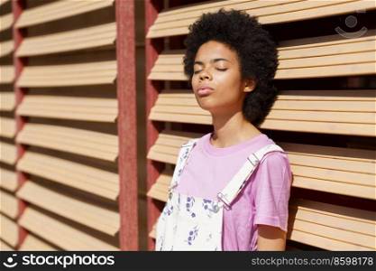 Calm African American female in casual outfit standing with closed eyes near wooden wall under bright sunshine on summer day. Tranquil black woman standing under sunlight