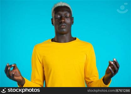 Calm african man in yellow relaxing, meditating. He calms down, breathes deeply with mudra om on blue studio background. Yoga concept. Calm african man in yellow relaxing, meditating. He calms down, breathes deeply with mudra om on blue studio background. Yoga concept.