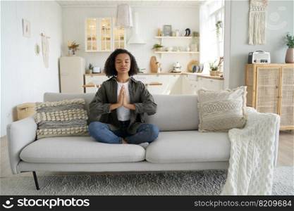 Calm biracial young teen girl girl practicing yoga, meditation at home. Serene mixed race woman meditates, sitting in lotus pose, relaxing on cozy couch. Healthy lifestyle, wellness concept.. Mixed race girl practices yoga, meditation at home sitting in lotus pose on couch. Healthy lifestyle