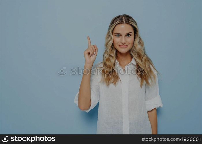 Calm blonde girl with slight smile pointing up in air with her finger, has new brilliant idea and showing it with gesture while standing next to blue wall. Female showing at copy space. Calm blonde girl with slight smile pointing up in air with her finger
