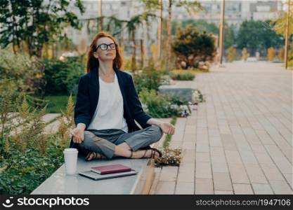 Calm business woman meditating in lotus outdoors, peaceful female freelance employee practicing yoga exercises while sitting in park after hard working day in office, holding hands in mudra. Calm business woman meditating in lotus outdoors, peaceful female freelance employee practicing yoga