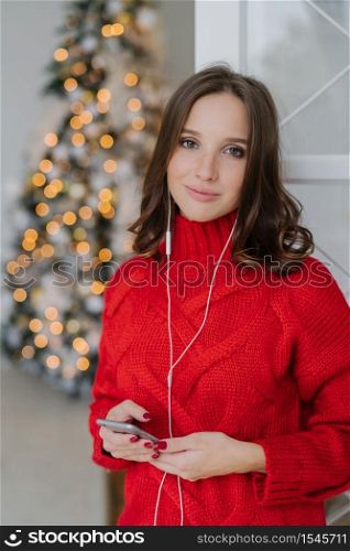 Calm carefree charming woman with healthy skin, dressed in red sweater, uses mobile phone and earphones, enjoys domestic atmosphere, spends Christmas time at home. People and technology concept