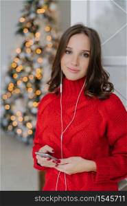 Calm carefree charming woman with healthy skin, dressed in red sweater, uses mobile phone and earphones, enjoys domestic atmosphere, spends Christmas time at home. People and technology concept