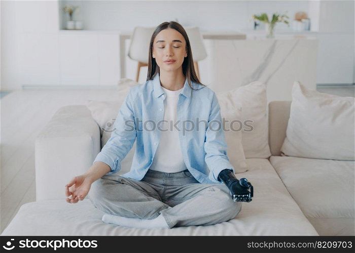 Calm disabled girl practicing yoga, sitting in lotus pose while meditation. Young female does mudra gesture with bionic prosthesis arm on sofa at home. Healthy lifestyle of people with disabilities.. Calm disabled girl does yoga with bionic prosthesis arm at home. Healthy lifestyle and disability