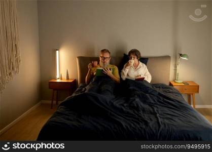 Calm family couple on retirement reading book together and drinking coffee while lying in bed before sleeping. Calm family couple on retirement reading book together in bed