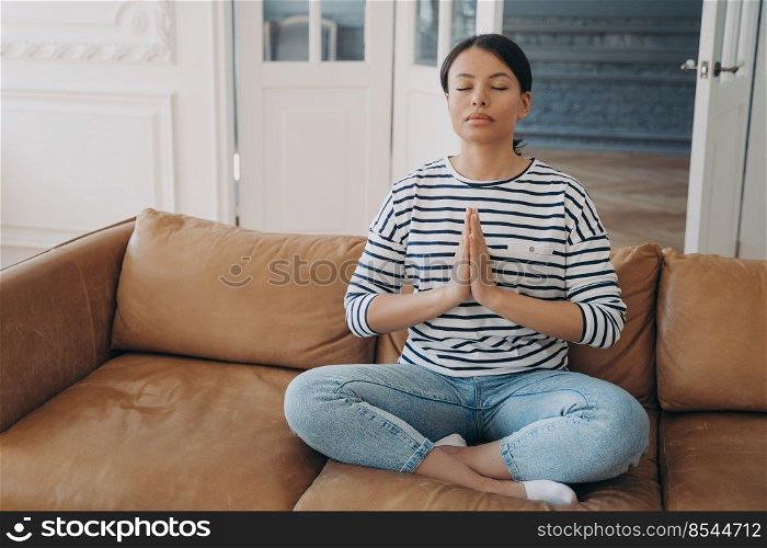 Calm female doing yoga exercises, sitting in lotus pose on sofa at home. Peaceful woman meditates folded hands in namaste gesture on couch in living room. Healthy lifestyle, mental health concept.. Calm female practices yoga sitting in lotus pose on sofa at home. Healthy lifestyle, meditation