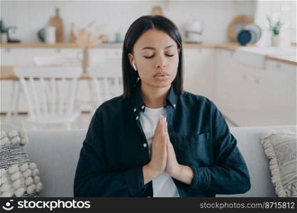 Calm female listening to meditation in earphone with namaste gesture to relieve stress at home. Young woman meditates, sitting on sofa with closed eyes, listens to relaxing music. Healthy lifestyle.. Calm female listening to meditation in earphone with namaste gesture to relieve stress at home