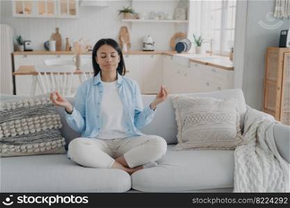 Calm female practice yoga, meditating, sitting on couch in lotus position with closed eyes at home. Serene woman does breathing exercises for mental balance. Healthy lifestyle, anxiety relief.. Calm female practice yoga, meditating, sitting on couch in lotus position at home. Healthy lifestyle
