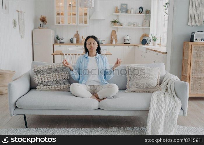Calm female practices yoga, breathing, relaxing on modern sofa at home. Young woman meditates, sitting on comfortable couch in cozy interior. Healthy lifestyle, emotion management, stress relief.. Female practices yoga, breathing, relaxing on cozy couch at home. Healthy lifestyle, stress relief