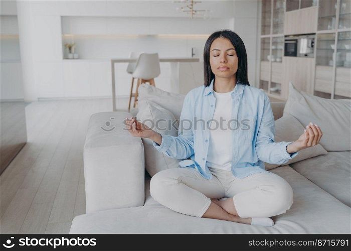 Calm female practicing yoga, sitting in lotus pose on sofa. Serene young woman keeping calmness, meditating on couch at home, reducing stress. Meditation, healthy lifestyle concept.. Female practicing yoga, sitting in lotus pose on sofa at home. Meditation, healthy lifestyle