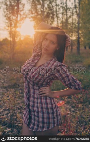 Calm girl posing in woodland park in autumnal time at sunset dressed in checkered shirt.