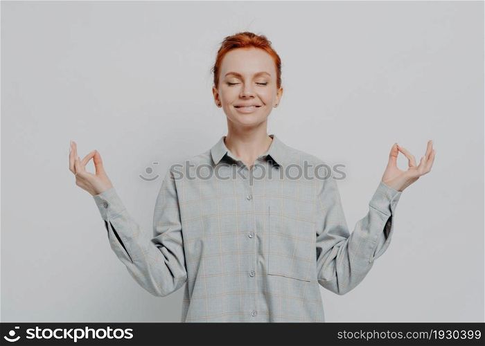 Calm happy 30s redhaired female in checkered shirt meditating with hands in mudra gesture and eyes closed, isolated on grey background, smiling ginger girl controling controling negative emotions. Calm redhaired female in checkered shirt meditating with hands in mudra gesture and eyes closed