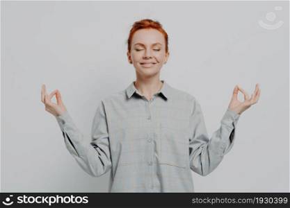 Calm happy 30s redhaired female in checkered shirt meditating with hands in mudra gesture and eyes closed, isolated on grey background, smiling ginger girl controling controling negative emotions. Calm redhaired female in checkered shirt meditating with hands in mudra gesture and eyes closed