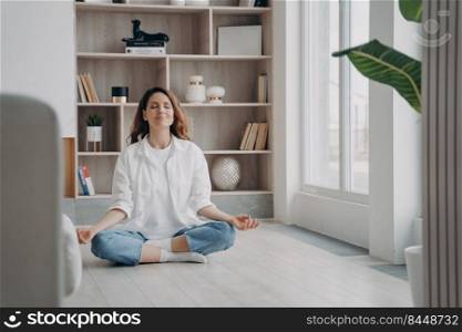 Calm happy latin woman is practicing yoga and meditation on floor at home. Girl is sitting near window in morning in lotus asana. Morning gymnastics. Stress relief and mind balance concept.. Calm happy latin woman is practicing yoga. Girl is sitting near window in morning in lotus asana.