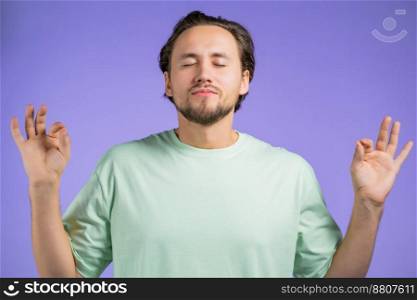 Calm man in basic t-shirt relaxing, meditating. He calms down, breathes deeply with mudra om on violet studio background. Yoga concept. Calm man in basic t-shirt relaxing, meditating. He calms down, breathes deeply with mudra om on violet studio background. Yoga concept.