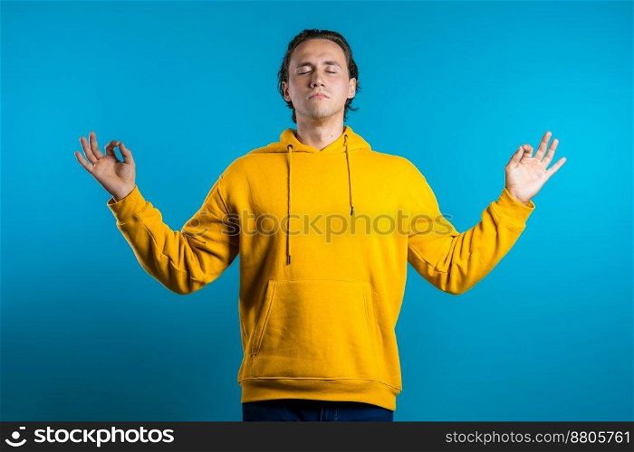 Calm man in yellow wear relaxing, meditating. He calms down, breathes deeply with mudra om on blue studio background. Yoga concept. High quality photo. Calm man in yellow wear relaxing, meditating. He calms down, breathes deeply with mudra om on blue studio background. Yoga concept.