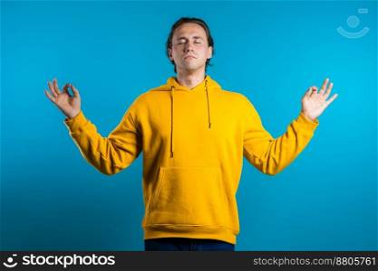 Calm man in yellow wear relaxing, meditating. He calms down, breathes deeply with mudra om on blue studio background. Yoga concept. High quality photo. Calm man in yellow wear relaxing, meditating. He calms down, breathes deeply with mudra om on blue studio background. Yoga concept.