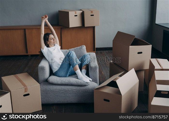 Calm satisfied young woman resting on armchair after packing cardboard boxes for moving in≠w apartment. Hispanic girl stretχng arms, dreaming about future home∫erior. Relocation, first rea<y.. Happy woman rests after packing boxes for moving, dreams about future home. Relocation, first rea<y