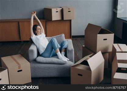 Calm satisfied young woman resting on armchair after packing cardboard boxes for moving in≠w apartment. Hispanic girl stretχng arms, dreaming about future home∫erior. Relocation, first rea<y.. Happy woman rests after packing boxes for moving, dreams about future home. Relocation, first rea<y