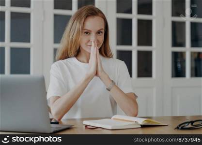 Calm smiling woman joins hands in prayer gesture, hopes for success of her business, wishes dreams come true, visualizes the future. Hopeful female employee sitting at desk with laptop, dreaming.. Calm female joins hands in prayer gesture hopes for success, believes in dream, sitting laptop