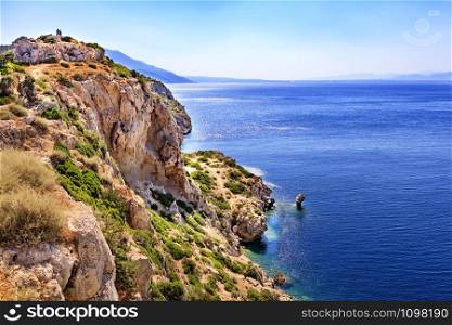 Calm turquoise waves wash the rocky shore of the Gulf of Corinth in the Ionian Sea in the rays of the dazzling and hot sun.. Seascape from the rocky steep coast of the Ionian Sea in Greece.
