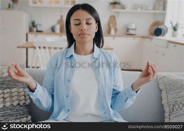 Calm woman meditate folded fingers in mudra hand gesture sitting on sofa at home. Peaceful young female practice yoga, relieve negative emotions, relax on couch in living room. Emotion management.. Calm woman practices yoga meditating folded fingers in mudra hand gesture sitting on sofa at home