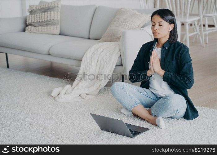 Calm woman meditates to relieve stress, practicing yoga at laptop in lotus asana on floor at home. Female in earphones feels calmness, listening audio affirmations, meditation. Healthy lifestyle.. Calm woman meditates to relieve stress, practicing yoga at laptop in lotus asana on floor at home