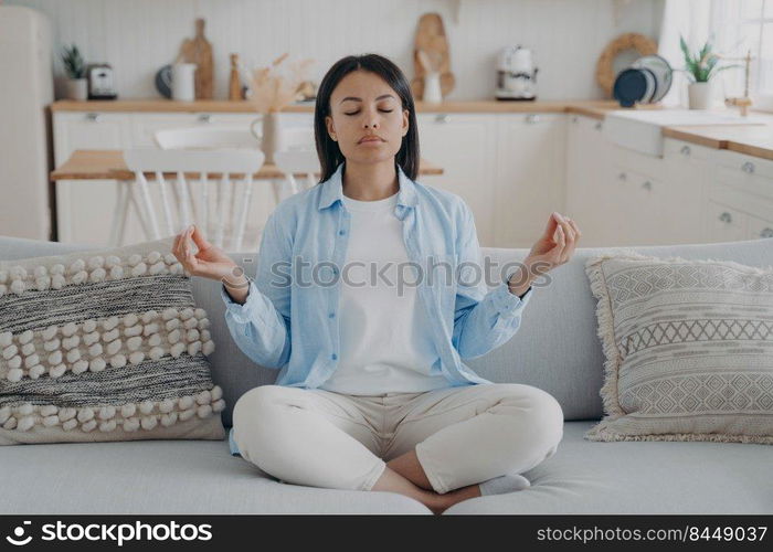 Calm woman practice yoga, sitting on sofa in lotus position, eyes closed, enjoy meditation at home. Female meditate, breathing for mental balance, relaxing. Healthy lifestyle, anxiety relief concept. . Calm woman meditate, practice yoga, sitting on sofa in lotus position at home. Healthy lifestyle