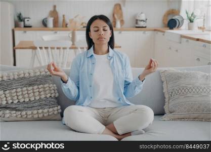 Calm woman practice yoga, sitting on sofa in lotus position, eyes closed, enjoy meditation at home. Female meditate, breathing for mental balance, relaxing. Healthy lifestyle, anxiety relief concept. . Calm woman meditate, practice yoga, sitting on sofa in lotus position at home. Healthy lifestyle