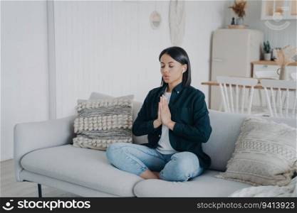 Calm woman practices yoga, meditates in lotus pose on couch. Namaste gesture, relaxation at home. Healthy lifestyle, stress relief.