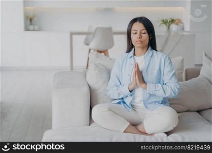 Calm woman practices yoga, meditates, sitting in lotus pose on sofa at home. Female sits on cozy couch relaxing folded hands in namaste gesture. Wellness, stress relief, healthy lifestyle concept.. Calm woman practices yoga, meditates, sitting in lotus pose on sofa at home. Wellness, stress relief