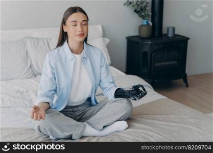 Calm young disabled girl with bionic hand prosthesis practicing yoga sitting on bed at home. Female with artificial arm limb meditates folded fingers in mudra gesture. Disability and healthy lifestyle. Disabled girl with bionic prosthetic hand does yoga on bed at home. Disability and healthy lifestyle
