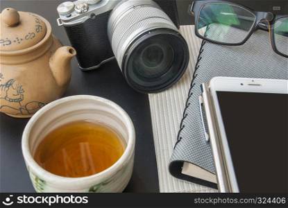 Camera, glasses, mobile phones, car keys, pens, 2 notebooks and Chinese tea placed on a black table during break from work