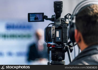 Cameraman wearing headphones and recording male speaker standing on the stage at press conference media event.