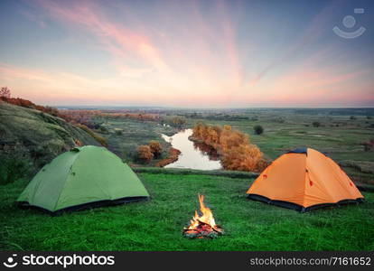 Camping from the orange and green tents with a fire on the banks of the river. Early morning. Summer landscape. concept of travel and solitude. Camping from orange and green tents with fire on banks of river