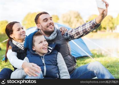 camping, hike, technology and people concept - happy family with smartphone taking selfie at campsite