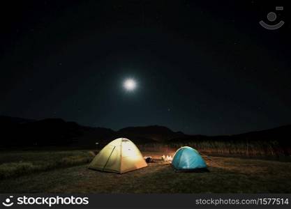 Camping in the mountains under the moon. A tent pitched up and glowing under the sky.. Tourist tents in camp among meadow in the night mountains