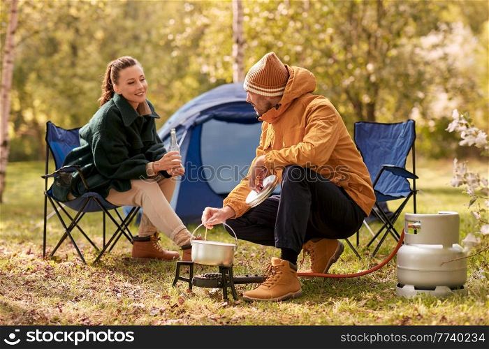 camping, tourism and travel concept - happy couple drinking beer and cooking food in pot on tourist gas burner at tent camp. couple drinking beer and cooking food at tent camp