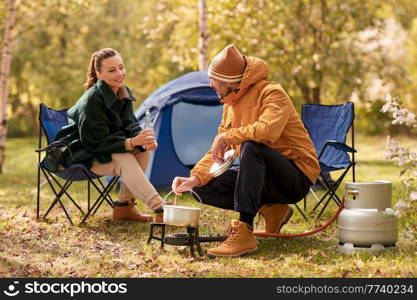 camping, tourism and travel concept - happy couple drinking beer and cooking food in pot on tourist gas burner at tent camp. couple drinking beer and cooking food at tent camp