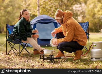 camping, tourism and travel concept - happy couple drinking beer and cooking food in pot on tourist gas burner at tent camp. couple drinking beer and cooking food at tent camp