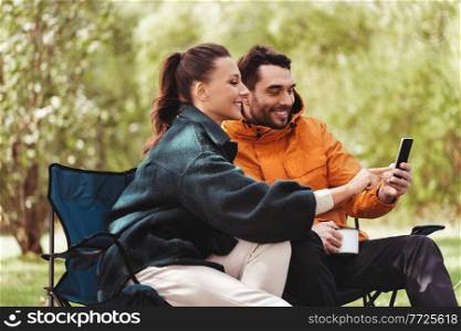 camping, tourism and travel concept - happy couple with smartphone drinking tea at campsite. couple with smartphone drinking tea at campsite