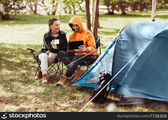camping, tourism and travel concept - happy couple with tablet pc computer drinking tea at tent camp. couple with tablet pc drinking tea at tent camp