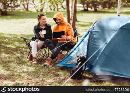 camping, tourism and travel concept - happy couple with tablet pc computer drinking tea at tent camp. couple with tablet pc drinking tea at tent camp