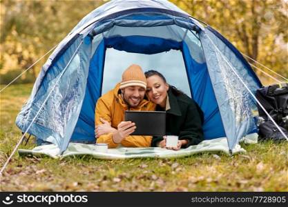 camping, tourism and travel concept - happy couple with tablet pc computer lying inside tent and drinking tea at campsite. couple with tablet pc drinking tea inside tent